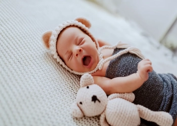 Side view of adorable newborn baby wrapped in wool scarf and with little cap on head while lying next to toy on fur blanket.