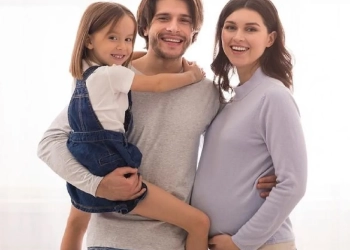 Portrait of cheerful pregnant mother, father and their little daughter posing together against window at home