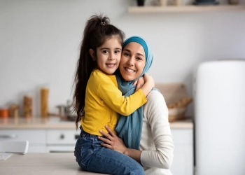 Portrait Of Happy Muslim Mother In Hijab And Little Daughter Posing In Kitchen Interior, Cute Small Girl Hugging Her Islamic Arab Mom And Smiling At Camera, Mommy And Child Having Fun At Home