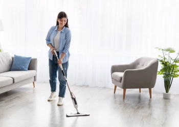 Portrait of casual woman washing hardwood laminate flooring using water spray mop pad and refillable bottle with cleaning agent. Chores Concept. Blurred Background, Free Copy Space, Modern Interior